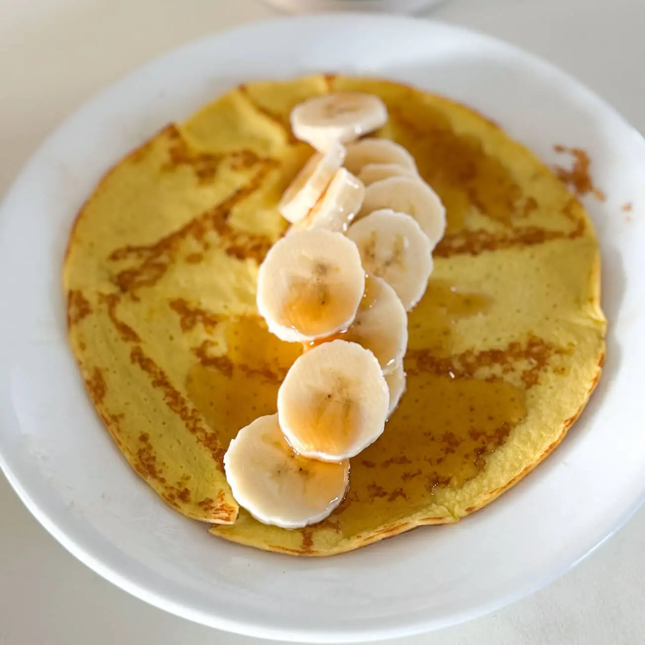 A plated high-protein pancake made with cottage cheese and eggs. It is garnished with fresh banana slices and maple syrup, styled with an organic syrup bottle slightly blurred in the background for depth.